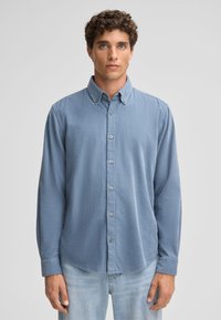 Lichtblauw button-up shirt met een kraag, lange mouwen en zichtbare knopen. De stof heeft een gladde textuur en wordt gedragen over lichte spijkerbroek.
