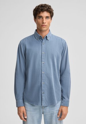 Light blue button-up shirt with a collar, long sleeves, and visible buttons. The fabric has a smooth texture, worn over light jeans.