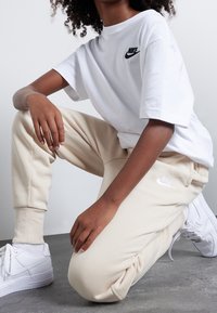 Bílý Nike tričko s černým logem, béžové tepláky s bílým logem a bílé tenisky. Hladká tkanina a uvolněný střih po celém obvodu.