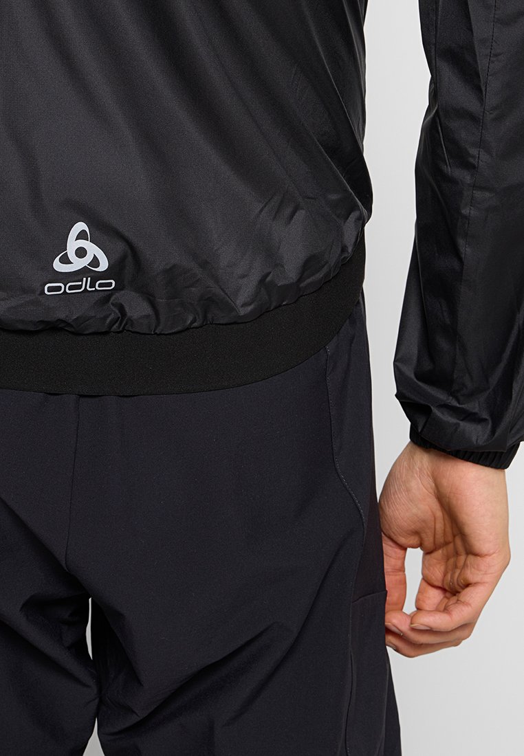 Czarna lekka kurtka z elastycznym dołem, z detalem logo i gładką fakturą, w połączeniu z ciemnymi spodniami.