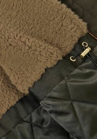 Brown fuzzy fabric and black quilted material overlap, featuring a gold hardware detail and a decorative drawstring for adjustment.