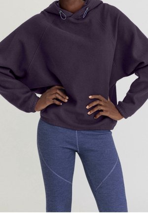 Person, die einen dunkelvioletten Kapuzen-Fleece mit Kordeln und eng anliegende blaue Leggings trägt, die Hände auf den Hüften vor einem schlichten Hintergrund.