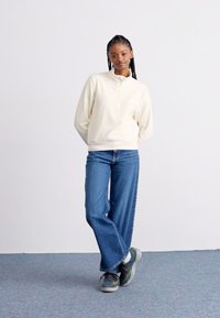 Lys beige zip-genser med lange ermer, parret med blå vide jeans og grå sneakers med grønne detaljer, mot en enkel bakgrunn.