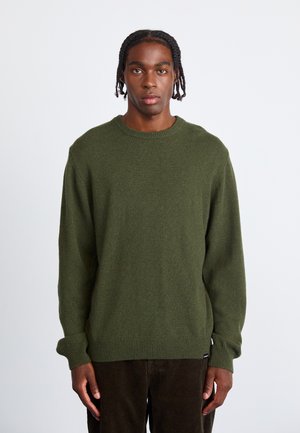 Young man with braided hair wearing an olive green knit sweater and dark brown corduroy pants, standing against a plain white background.