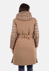 Cappotto lungo imbottito beige con cappuccio staccabile. Presenta una vita con cintura, tessuto liscio e maniche trapuntate con finitura lucida.