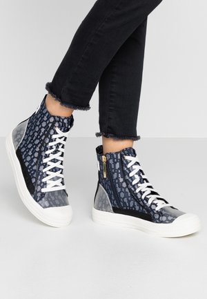 Personne portant un jean noir raccourci et des baskets montantes bleu marine à motifs avec des lacets blancs et des fermetures éclair latérales, debout sur un sol gris.