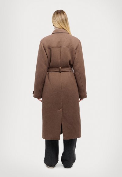 Bruine, lange jas met een gestructureerde stof, ceintuur in de taille en een opening achteraan. Heeft een kraag en een relaxed fit, gecombineerd met brede zwarte broek.