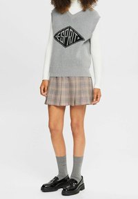 Jersey sin mangas gris con un logo de diamante negro, combinado con una falda plisada de cuadros que muestra tonos de gris, rosa y crema. Mocasines negros y calcetines grises.