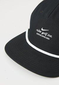 Căciulă de golf Nike neagră cu logo brodat alb, text "nike golf inc. ESTABLISHED 1984" și detaliu de frânghie albă deasupra borului.