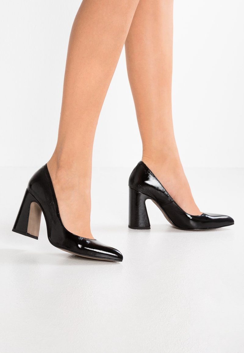 Escarpins en cuir verni noir à bout pointu et talon bloc épais. Ils présentent une texture lisse et brillante ainsi qu'une silhouette élégante.