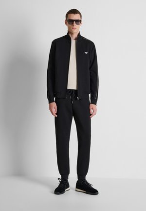 Hombre de pie junto a una pared blanca, con gafas de sol negras, chaqueta negra con cremallera, pantalones negros, camisa beige y zapatillas negras.