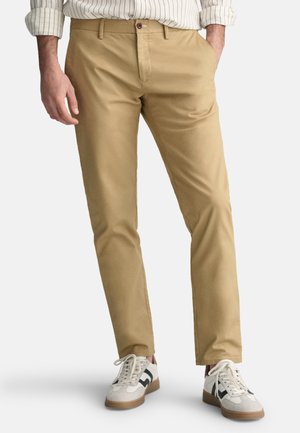 Tan chinos with a flat front, side pockets, and a slim fit. Worn with white sneakers featuring black accents. Cotton blend fabric.