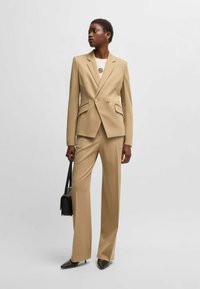 Vaalean beige räätälöity puku, jossa on tuplarivinen bleiseri ja leveälahkeiset housut. Silkkinen kangas ja terävät kaulukset. Yhdistetty valkoisen paidan ja mustan käsilaukun kanssa.