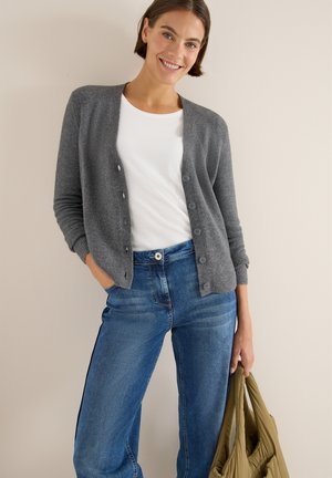 Grauer Strickcardigan mit rundem Ausschnitt, Knopfverschluss und gerippten Bündchen, getragen über einem weißen Hemd und kombiniert mit blauen Jeans. Hält eine grüne Tasche.