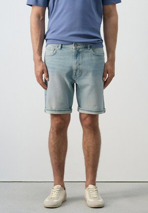 Hombre con pantalones cortos de mezclilla azul claro con dobladillo enrollado, camiseta azul y zapatillas blancas sucias, de pie sobre un suelo gris contra una pared lisa.