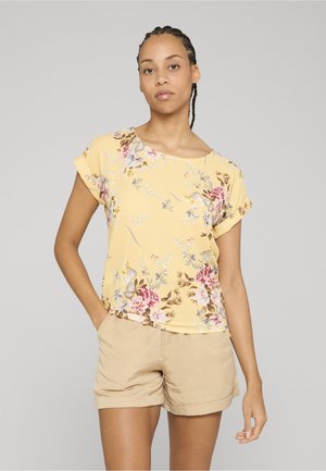 Femme aux cheveux tressés portant un haut court jaune à manches courtes à motif floral et un short beige à ourlet roulé, debout avec une main dans la poche.