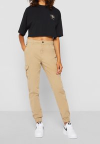 Personne portant un pantalon cargo jogging beige avec des chevilles élastiques, un t-shirt court noir, et des baskets blanches avec des logos Nike swoosh noirs.