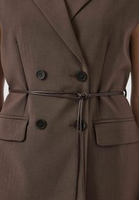 Bruine op maat gemaakte vest met een dubbelbreasted ontwerp, twee grote zwarte knopen, zijzakken en een dunne riem die de taille accentueert.