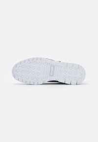 Chaussure de sport noire avec une semelle en caoutchouc texturée blanche, présentant des motifs géométriques et des détails de talon renforcés pour plus de durabilité.