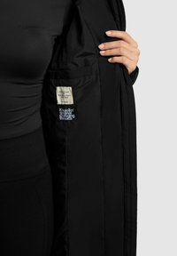 Schwarze gepolsterte Jackeninnenseite mit sichtbarem Etikett und Logo. Verfügt über eine glatte Textur, flache Nähte und eine mit Reißverschluss gesicherte Tasche.