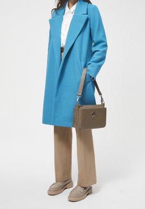 Blue wool coat with large collar, tan trousers, beige shoes, and a small brown handbag with a logo detail and adjustable strap.