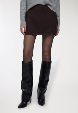 Donna che indossa stivali al ginocchio in pelle nera, collant neri trasparenti, mini gonna marrone scuro con spacco laterale e maglione grigio su sfondo bianco.