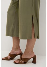 Olivgröna culotter med sidoklyftor, kombinerade med bruna klacksandaler med korsremmar och en kraftig klack. Slät tygstruktur.