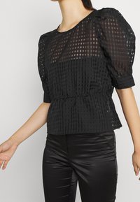 Top nero con maniche a palloncino e motivo a scacchi, realizzato in tessuto trasparente, con vita arricciata e scollatura tonda. Abbinato a pantaloni neri lucidi.