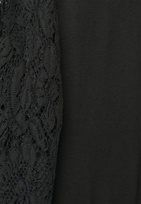 Melnas tekstila drēbes, kurām ir mežģīņu piedurkne ar lapu rakstu, kontrastējoši ar vienkāršu melnu audumu. Izteikta teksturēta mežģīņu detaļa.