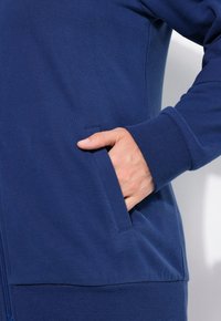 Felpa blu navy in tessuto, con zip frontale, polsini a coste e una tasca laterale dove è posizionata una mano. Texture morbida, design casual.