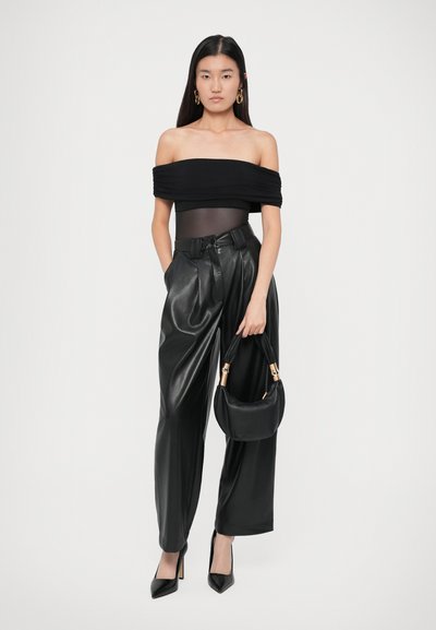 Sieviete ģērbusies melnā off-shoulder topā, augstās jostas augstuma platās ādas biksēs, melnās papēžkurpēs, tur rokās melnu rokassomu ar zelta rokturiem.