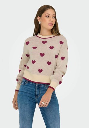 Vrouw in een beige gebreid trui met bordeauxrode harten, gecombineerd met een blauwe jeans, die naar de zijkant kijkt tegen een effen achtergrond.