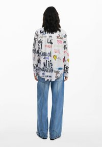 White button-up shirt featuring diverse text patterns in various colors, paired with wide-leg blue denim jeans. Text includes "LOVE" and "1984."