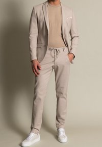 Beżowa marynarka dopasowana na beżowym T-shircie, w zestawie z pasującymi spodniami dresowymi i białymi sneakersami. Gładki materiał z minimalnymi detalami.