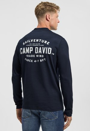 Mann mit blondem Haar, der ein marineblaues Langarmshirt mit weißem "Camp David Sailventure"-Schriftzug auf dem Rücken und blaue Jeans trägt.