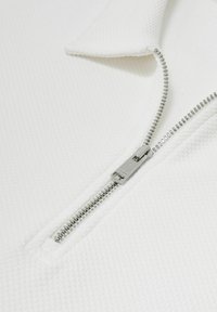 Nærbillede af en delvist åbnet sølvlynlås på tekstureret hvidt stof med en foldet kravekant.