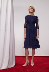 Robe de marine avec un bustier en dentelle et des manches trois-quarts, taille élastique et jupe fluide ; associée à des talons aiguilles de couleur claire.