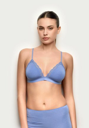 Donna con capelli lisci raccolti indossa un reggiseno azzurro chiaro con pizzo e slip coordinati a vita alta su uno sfondo semplice.