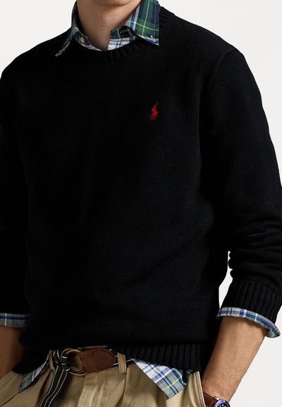 Suéter de malha preto com punhos e bainha canelados, apresentando um pequeno logotipo vermelho de polo. Usado sobre uma camisa xadrez azul e verde.