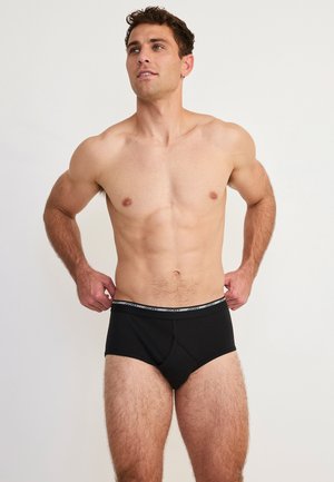 Atletyczny mężczyzna poprawia czarne bokserki z markowanym pasem, stoi na jasnym jednolitym tle, lekko spogląda w górę.