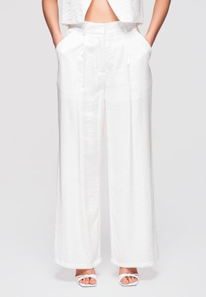 Femme portant un pantalon blanc taille haute à jambes larges, les mains dans les poches, et des sandales blanches à talons ouverts sur un fond neutre.