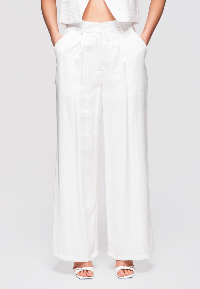Femme portant un pantalon blanc taille haute à jambes larges, les mains dans les poches, et des sandales blanches à talons ouverts sur un fond neutre.