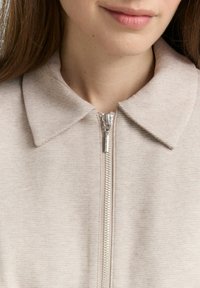 Lichtbeige zip-up jack met een kraaghalslijn, voorzien van een textuurstof en een zilveren ritsdetail aan de voorkant.