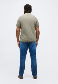 Άνδρας που στέκεται με την πλάτη στραμμένη φοράει χακί κοντομάνικο πουκάμισο, μπλε τζιν και καφέ παπούτσια μπροστά σε απλό λευκό φόντο.