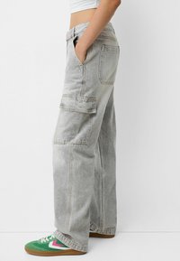 Pantalones cargo de denim gris holgados con bolsillos laterales, dobladillo deshilachado y lavado claro, combinados con zapatillas verdes y blancas con acentos rosas.