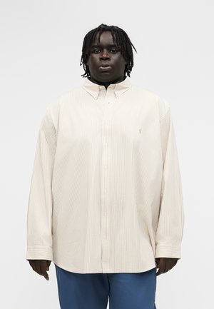 Bărbat cu piele închisă la culoare și dreadlocks scurți, purtând o cămașă bej supradimensionată cu dungi și pantaloni albaștri, pe un fundal simplu.