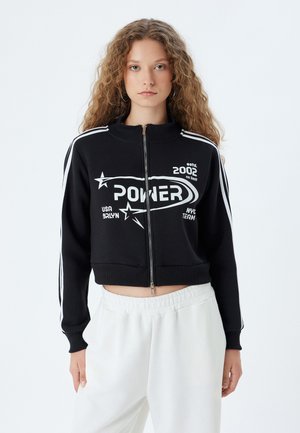 Sort cropped zip-up sweatshirt med hvide striber, der præsenterer "POWER" grafik og stardetaljer. Par med lyse bukser med elastisk talje.