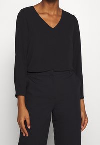 Blouse noire à manches longues avec un col en V, en tissu lisse, coupe décontractée, portée avec un pantalon sur mesure.