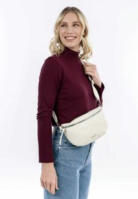 Sac en cuir blanc en forme courbée, avec une fermeture éclair argentée et une bandoulière ajustable, porté sur un top côtelé bordeaux avec un jean en denim.