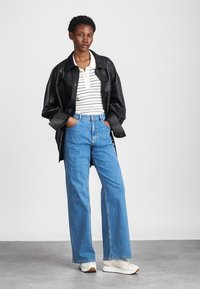Μαύρο δερμάτινο oversized μπουφάν, λευκή μπλούζα με ρίγες, ανοιχτό μπλε φαρδύ τζιν και λευκά αθλητικά παπούτσια. Λείες υφές, casual εφαρμογή.